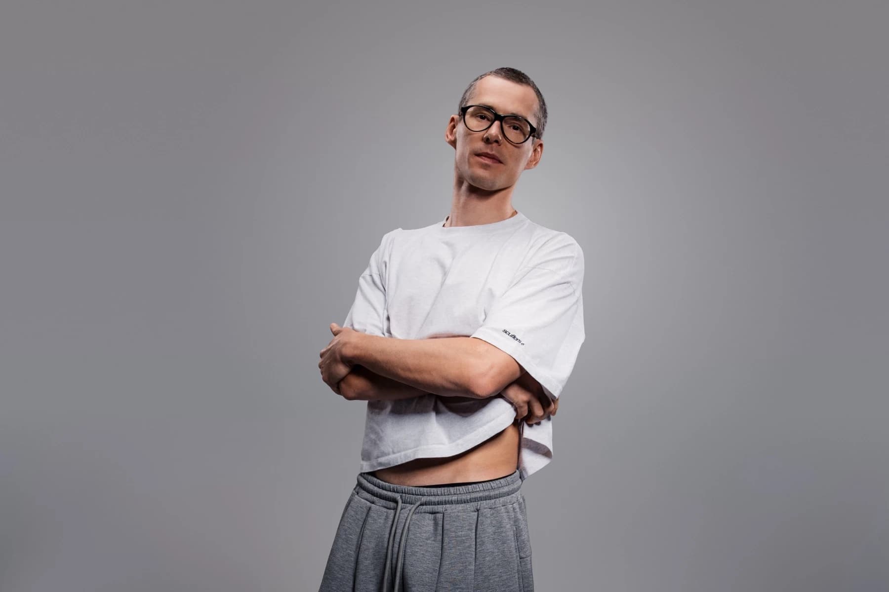 Lost Frequencies w okularach, białej koszulce i szarych spodniach dresowych, z założonymi rękami, na szarym tle studyjnym
