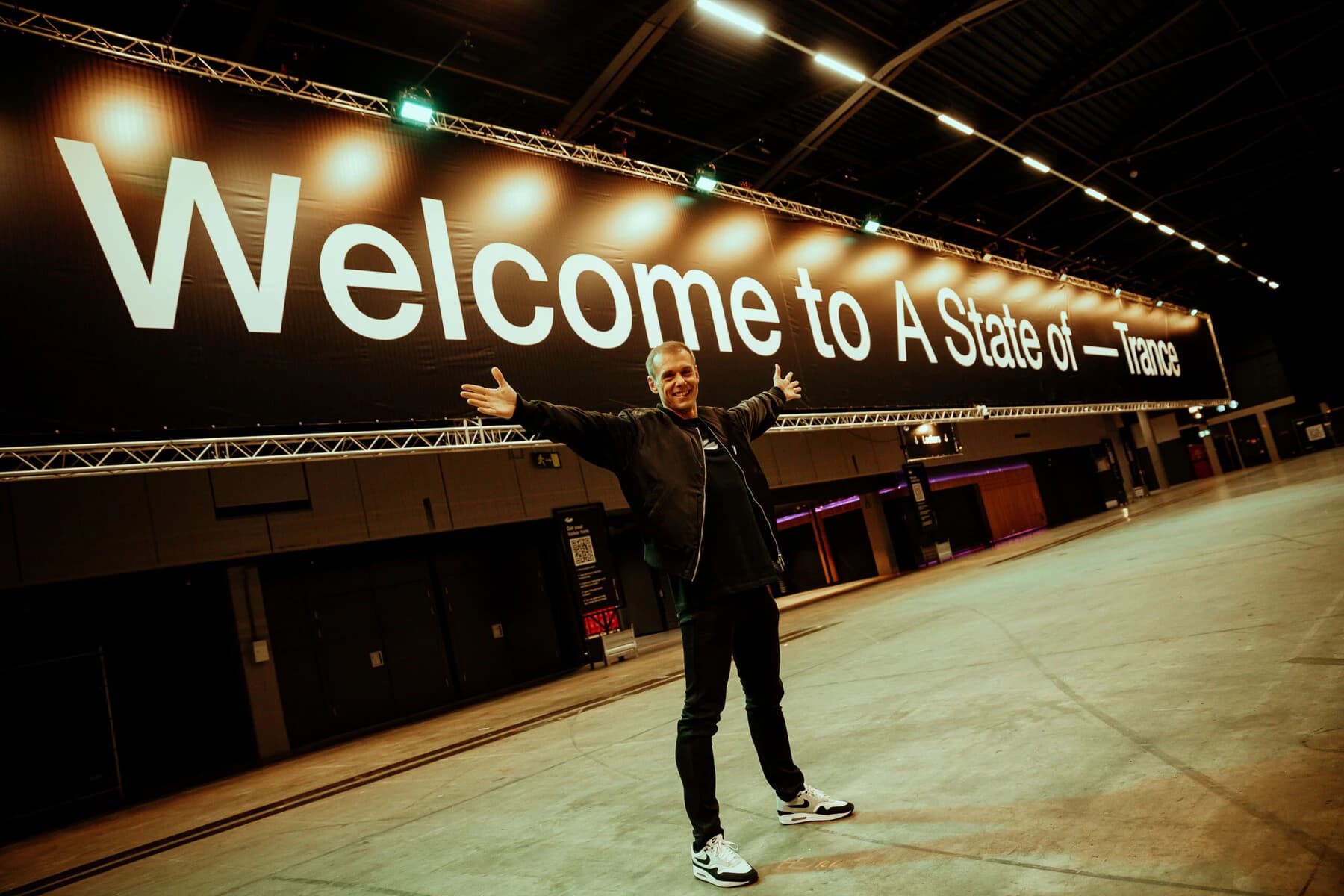 Armin van Buuren w czarnej kurtce, czarnych spodniach i białych sneakersach rozkłada ręce w pustej hali pod długim, podświetlonym reflektorami banerem z napisem „Welcome to A State of Trance”