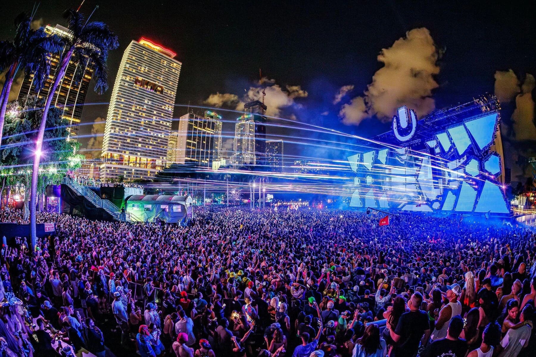 Główna scena Ultra Music Festival w Miami nocą: niebieska konstrukcja z podświetlonym logo „U”, smugi laserów nad gęstym tłumem, a po lewej palmy i oświetlone wieżowce