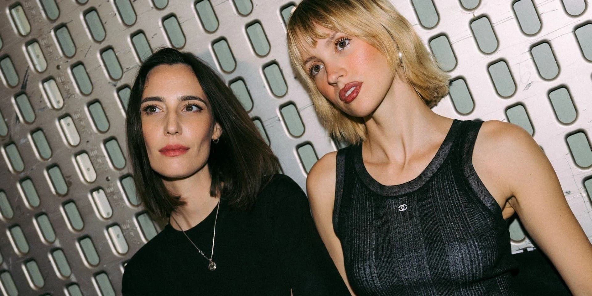 Amelie Lens w czarnej bluzce z ciemnymi włosami do ramion i Angèle w blond bobie z grzywką, w prążkowanym topie Chanel, pozują na tle srebrnej perforowanej ściany